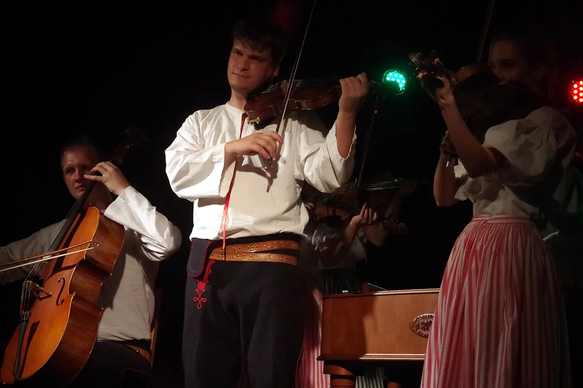 A violinist in a white shirt and embroidered trousers plays beside a bass player and a second violinist, with a cimbalom at the lower edge